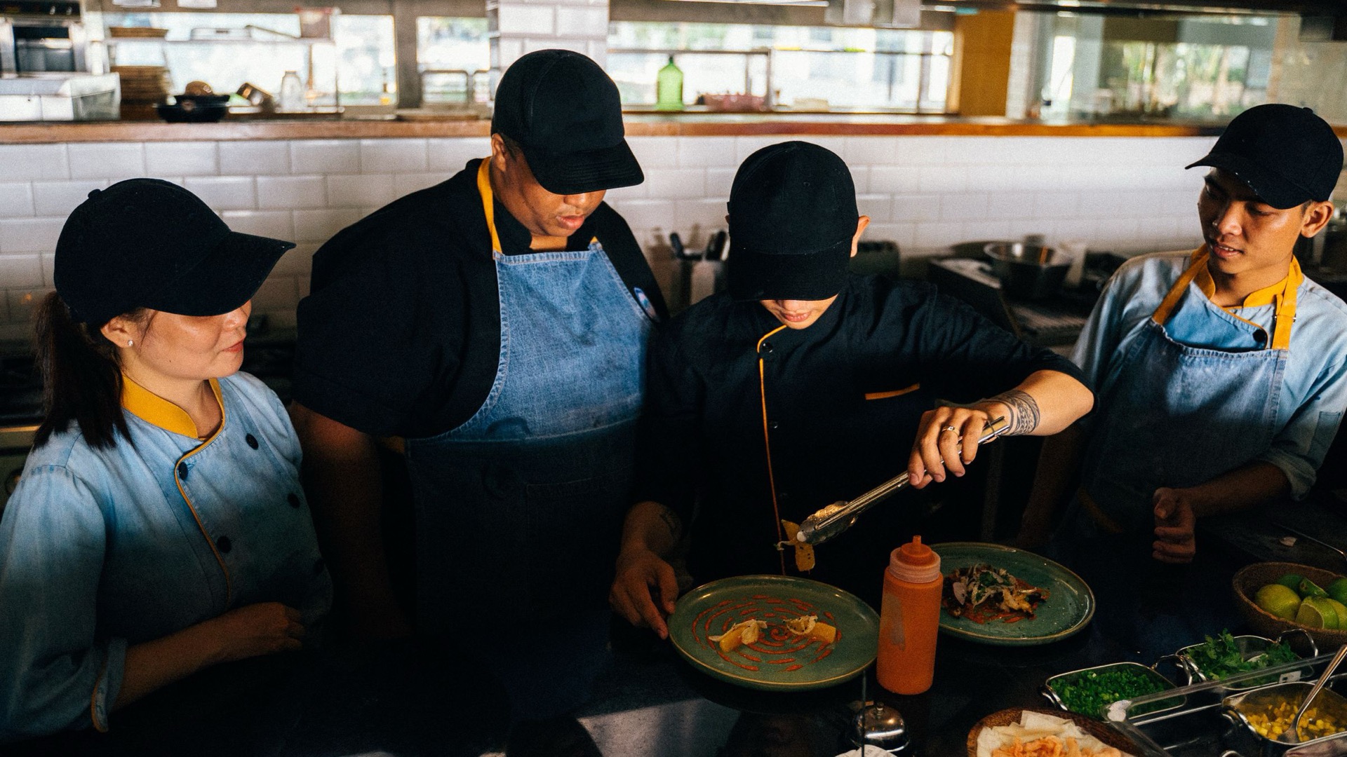 A head chef plating a dish while three cooks watch, a hands-on moment in a restaurant training program